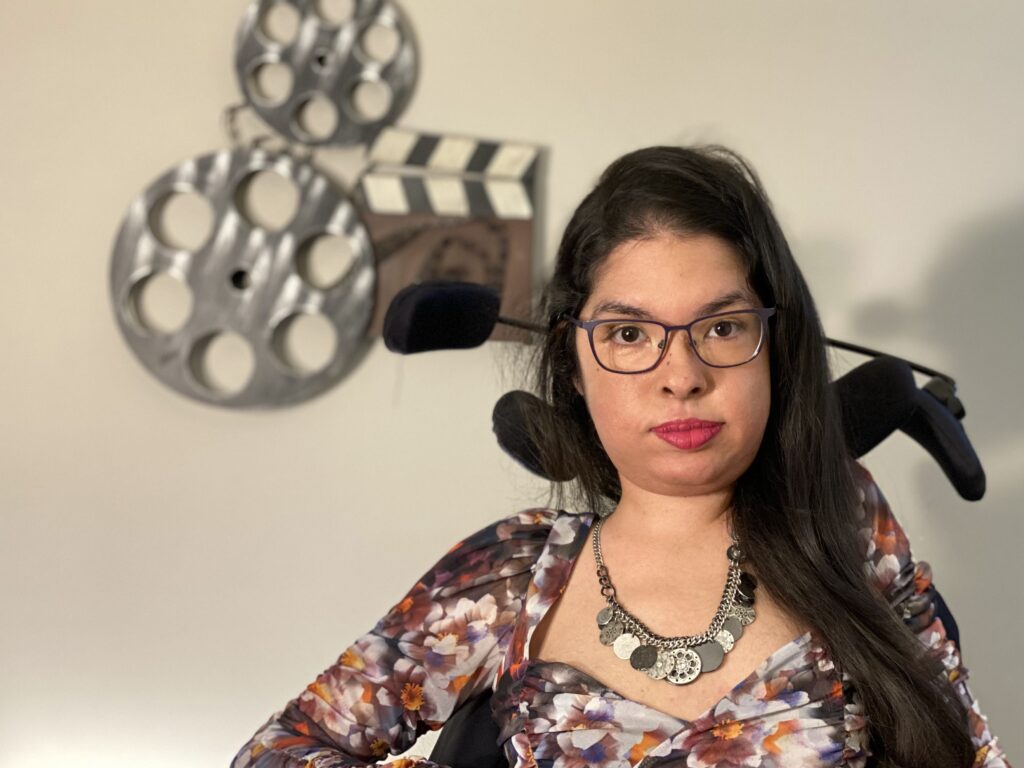 Michelle, a brown gal with long black hair in a floral top, poses in her power chair in front of a metal art piece featuring a slate and film reel.